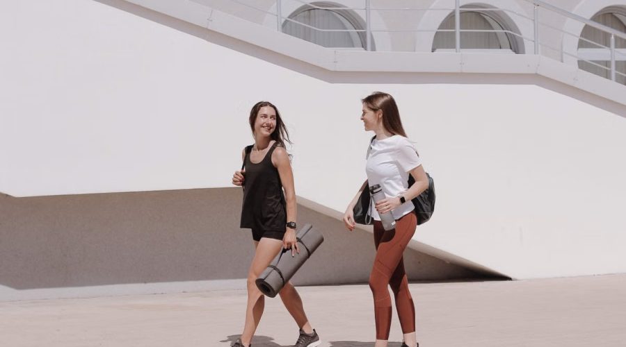 Two women in active wear walking outdoors while smiling and carrying yoga mats.