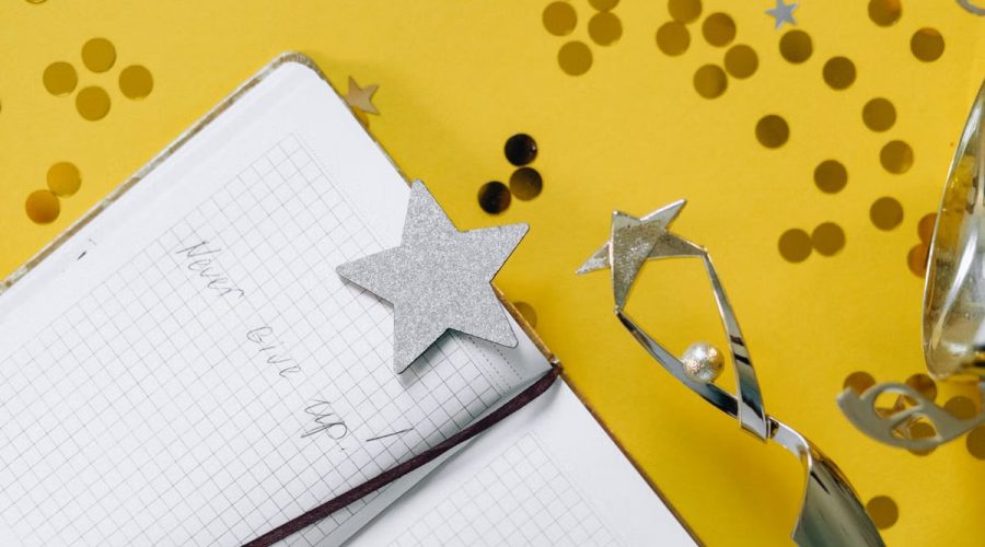 Flat lay of a notebook with motivational text, trophy, and star confetti on a yellow background.